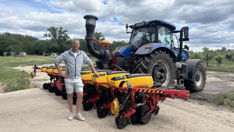 Egy férfi áll egy kék traktor és egy sárga vetőgép mellett egy mezőgazdasági területen.