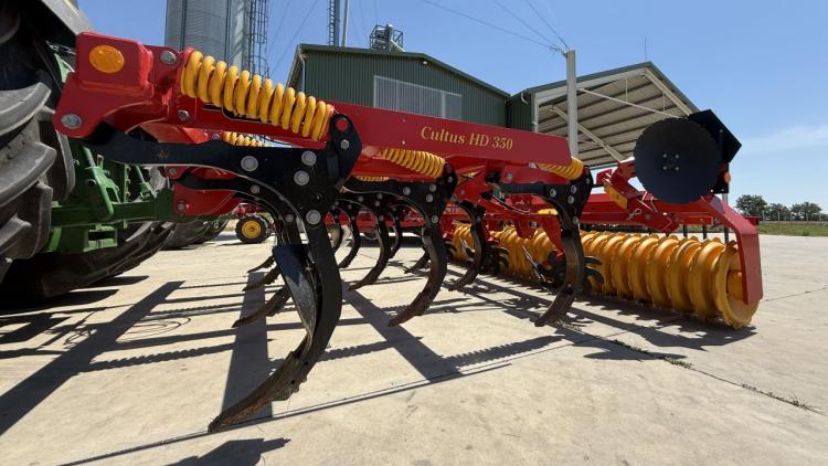 Egy piros és sárga mezőgazdasági gép, egy Cultus HD 350 talajművelő látható egy traktor mellett, egy farmudvaron. Egy piros és sárga mezőgazdasági gép, egy Cultus HD 350 talajművelő látható egy traktor mellett, egy farmudvaron.