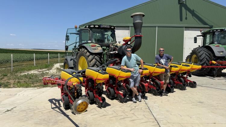 Két férfi áll egy nagy traktor és egy vetőgép mellett egy farm udvarán, kukoricafölddel a háttérben. Két férfi áll egy nagy traktor és egy vetőgép mellett egy farm udvarán, kukoricafölddel a háttérben.
